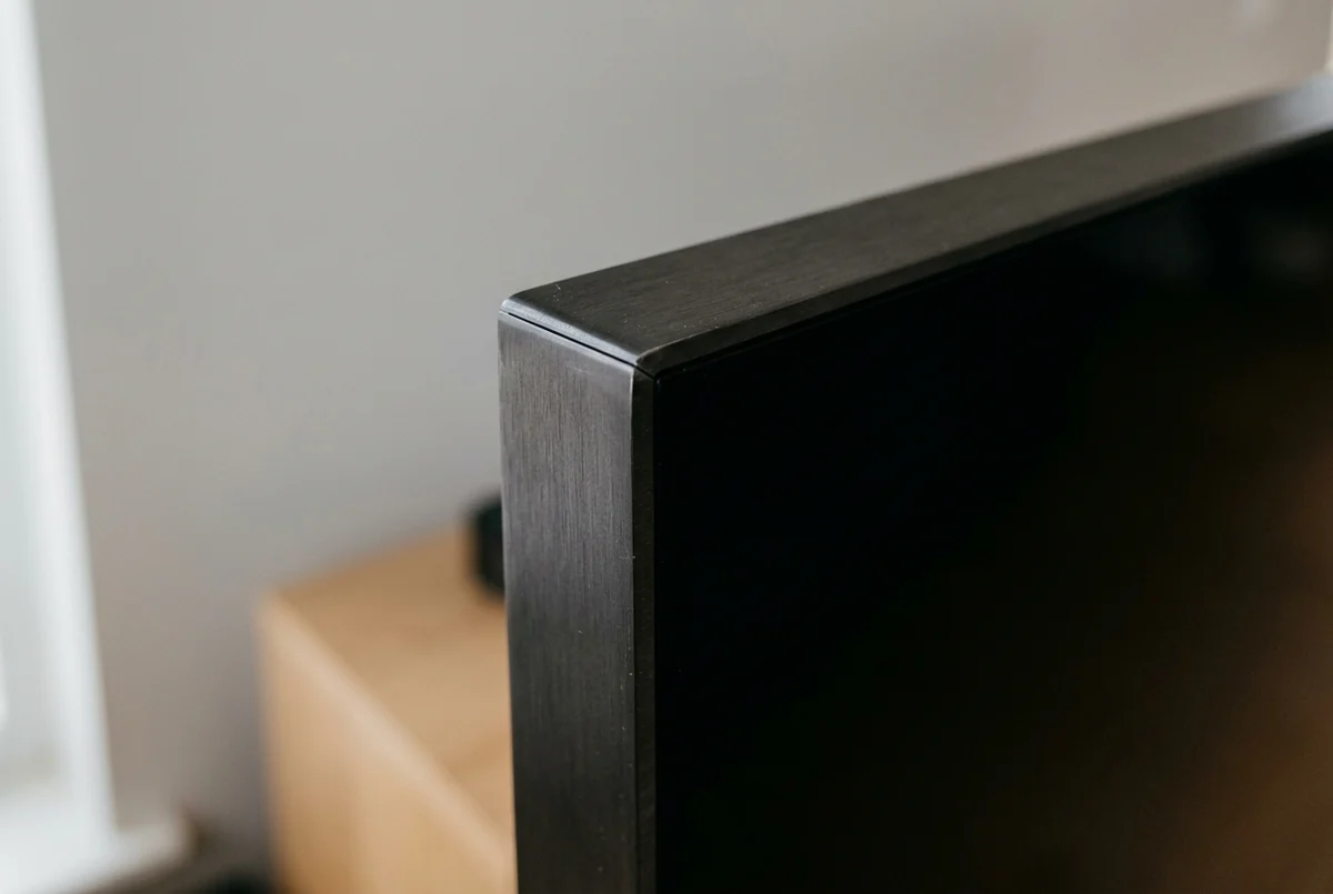 Canto da moldura preta de uma TV desligada, em close, com o rack de madeira desfocado ao fundo