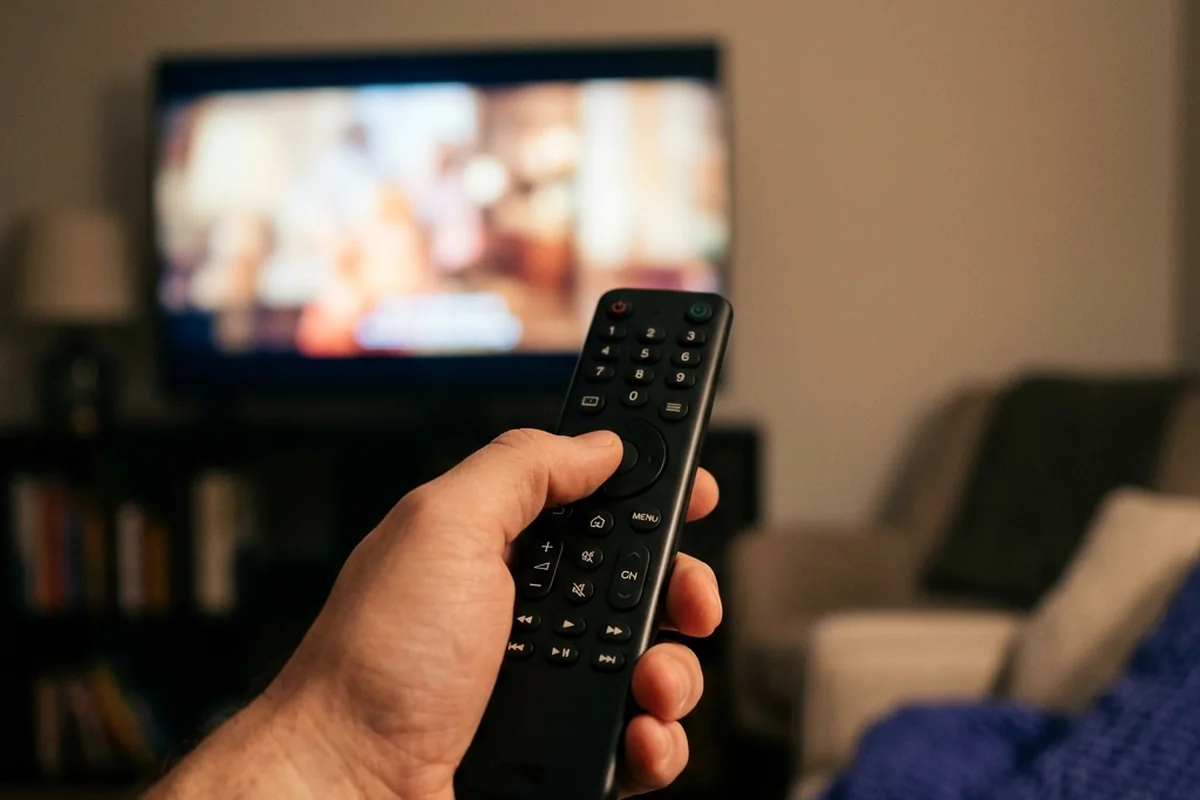 Mão segurando o controle remoto em uma sala escura, com televisão desfocada ao fundo.